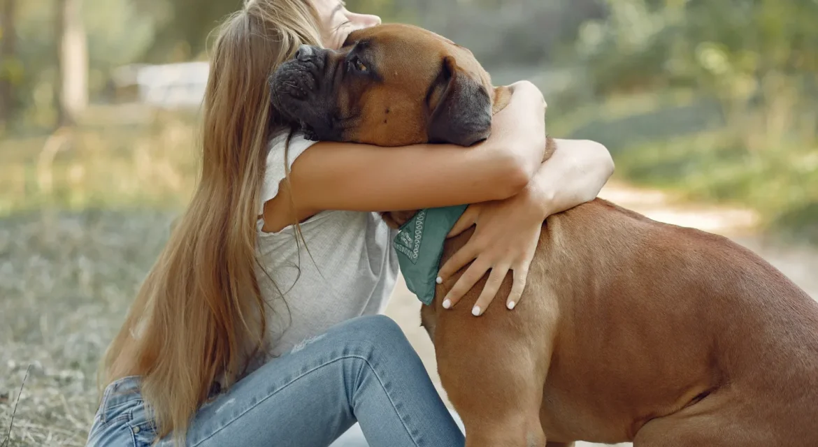 Una mujer abraza cariñosamente a un perro grande en un entorno natural, mostrando la conexión y el amor entre mascotas y humanos. La mujer viste una camiseta blanca y jeans, mientras el perro lleva un pañuelo azul.